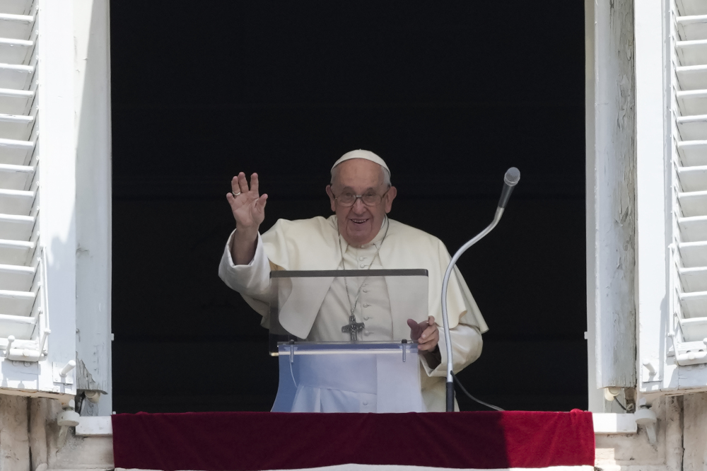 Pope Francis lifts his hand as he stands in his apartment window behind a clear lectern and microphone