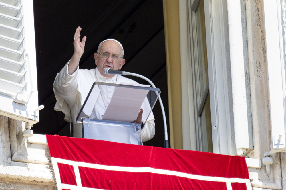 Pope Francis stands in his apartment window and raises his right hand as he speaks into a microphone
