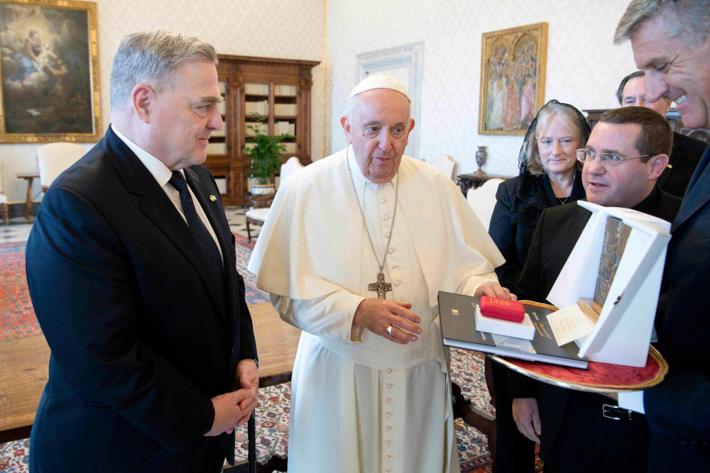 An older white man in a suit stands next to Pope Francis, who places his hand on an artwork another man is holding towards the first man
