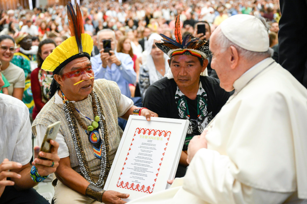 Two men wearing feather headpieces, one with a nose piercing, hand Pope Francis a frame with language