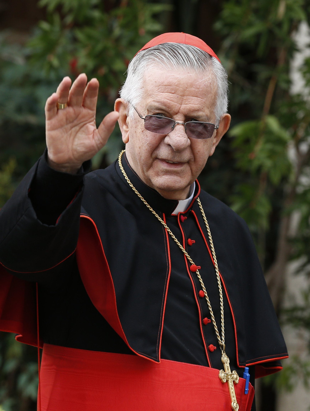 An older white man wearing cardinal's clothes and glasses transitioned to sunglasses raises his hand to wave.