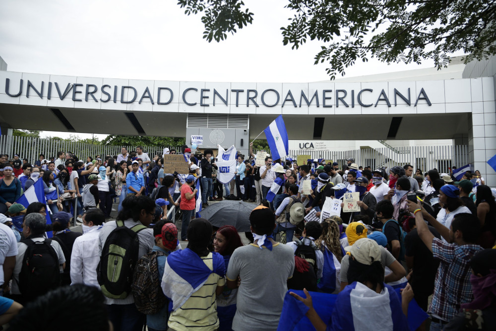 A group of people carrying Nicaraguan flags stand in front of an arch that reads "Universidad Centroamericana"