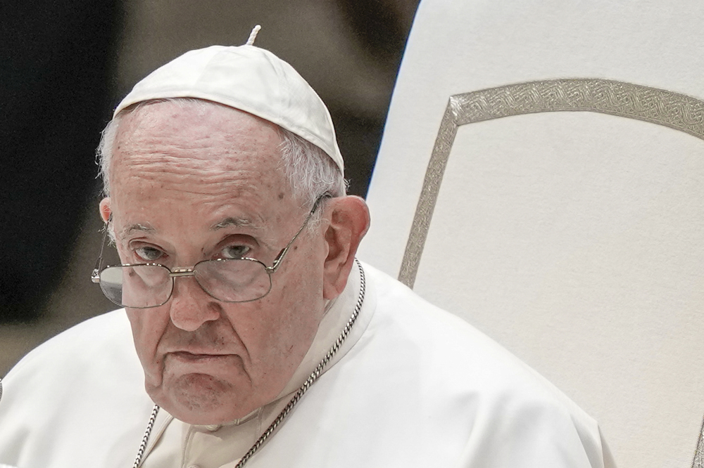 Pope Francis' head and shoulders are seen against a white chair. He wears glasses.