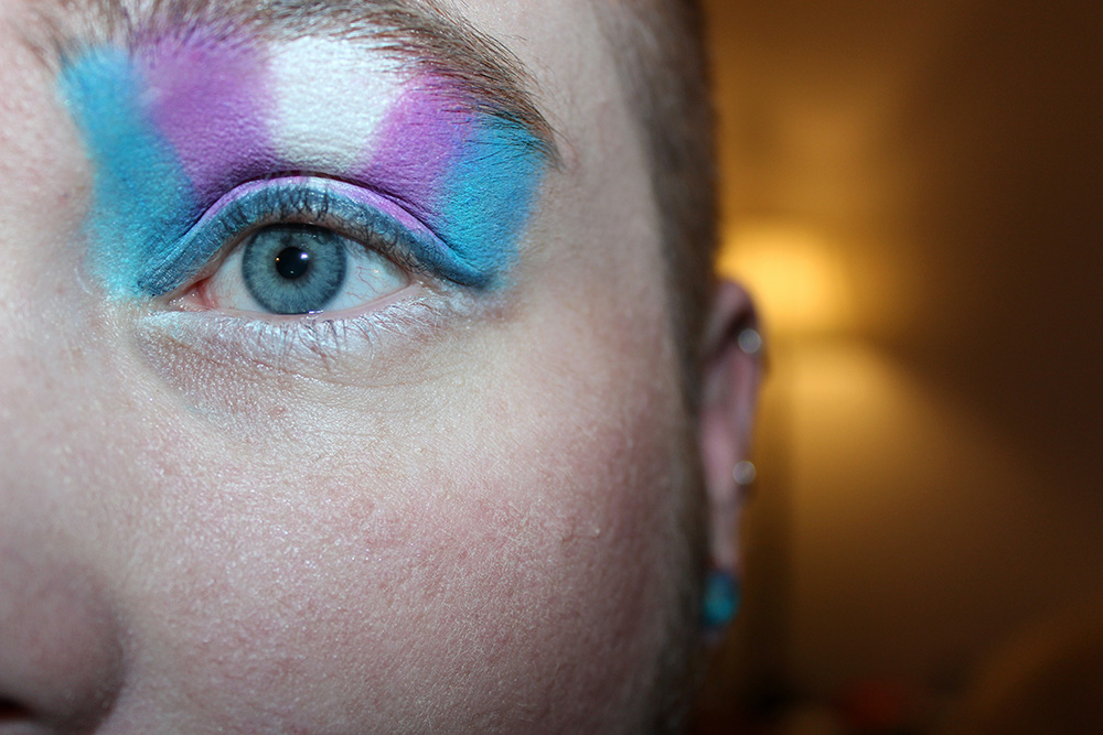 A blue eye with eye shadow in the colors of the trans flag (Unsplash/Kyle)