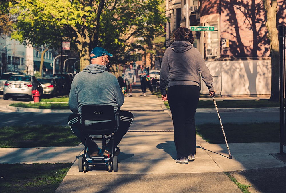 A person using a motorized scooter and a person using a cane proceed down a city sidewalk (Unsplash/Max Bender)