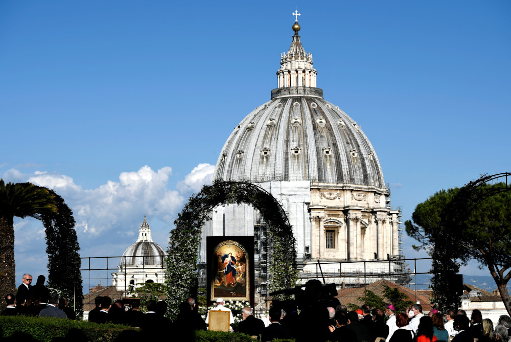 Leafy green arches are visible in front of the dome of St. Peter's Basilica with people sitting in the garden in front of a painting