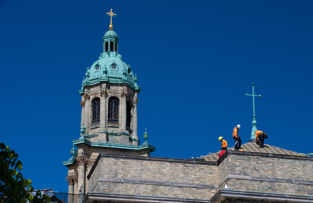 Construction workers stand on the roof of a church with a coppery green dome and cross