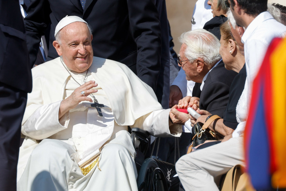 Pope Francis waves his hand while sitting in his wheelchair. With his other hand, he holds the hand of an older man in the crowd to the side.