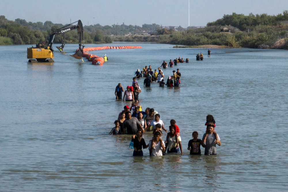 People, many with children on their shoulders, walk out into the middle of a river. A construction machine and buoys can be seen behind them.