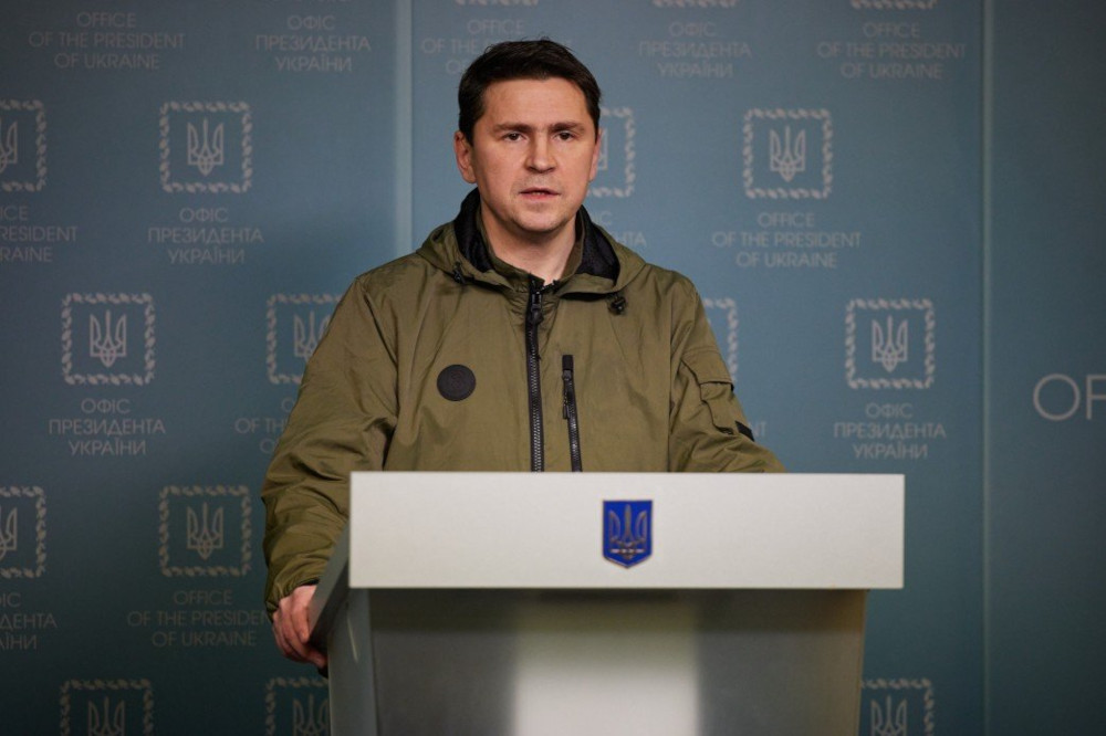 A white man with short dark hair wearing a green jacket speaks at a podium