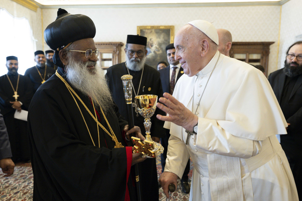 Pope Francis smiles at an older brown man with a beard wearing a black hat in a Hershey kiss shape, black robes, and long necklaces.