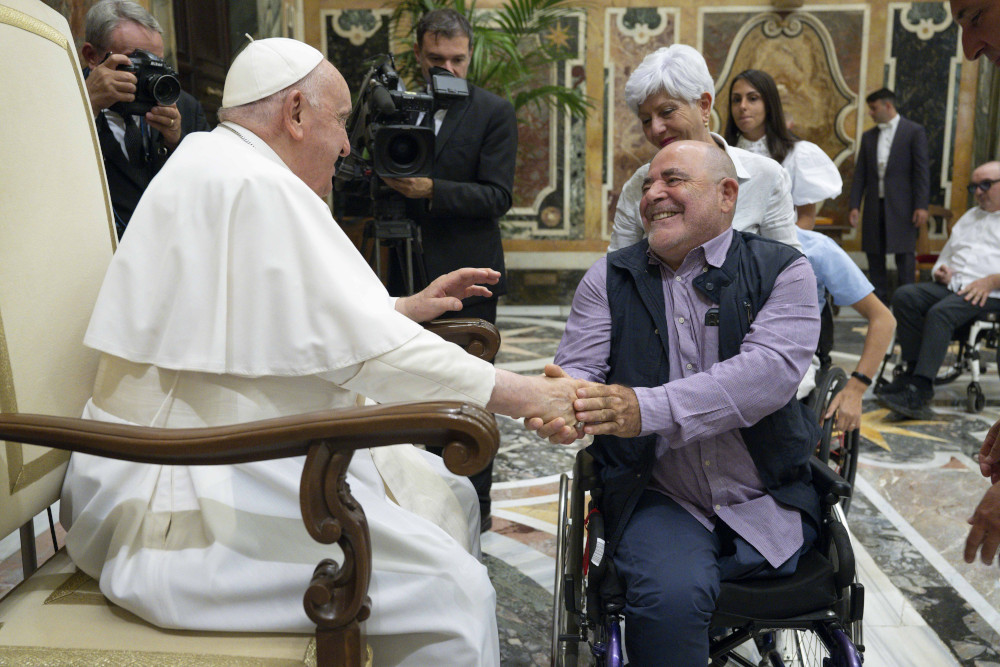 Pope Francis, sitting in a chair, shakes the hand of a man using a wheelchair
