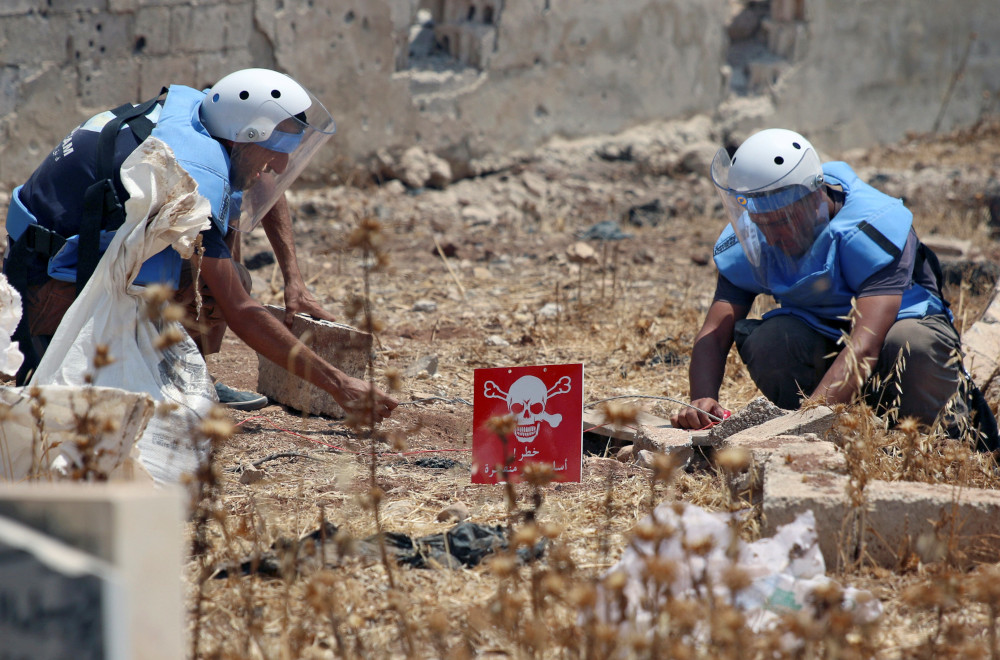 Two people wearing white helmets and light blue jackets crouch on the ground next to a red sign with a skull and crossbones