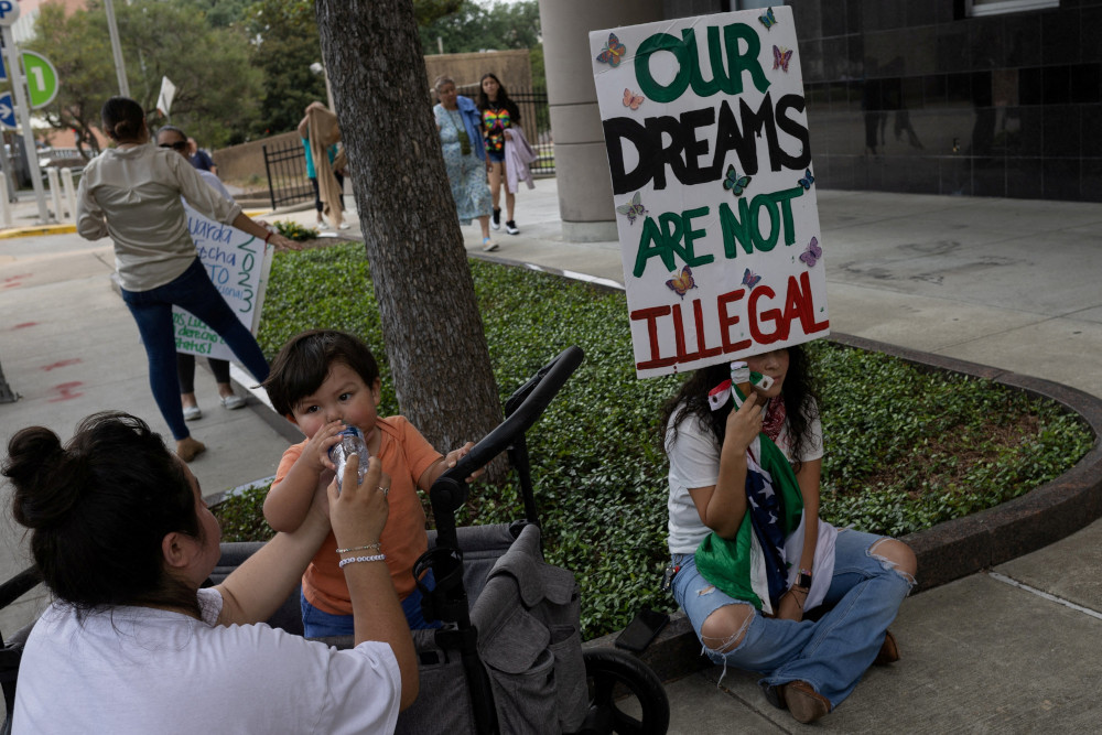 A person sitting cross-legged holds a sign that says "Our dreams are not illegal," while next to a person feeding a toddler.