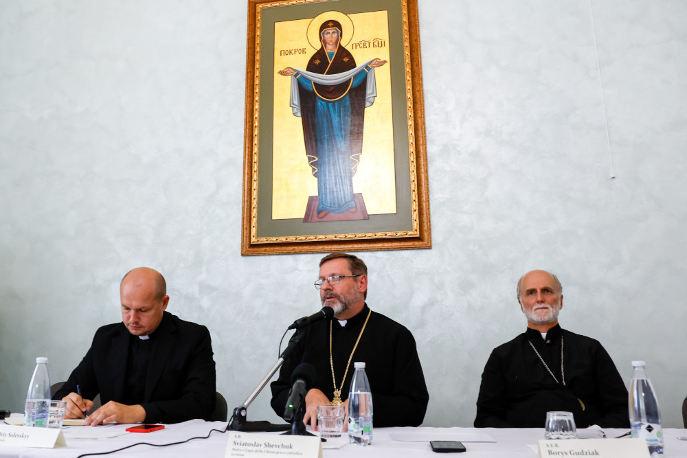 Three men wearing clerical collars sit at a table below a painting of a woman saint