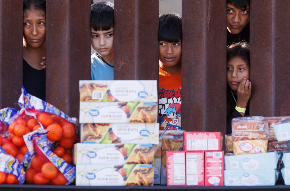 Brown people of different ages peer between the brown columns of the border wall at stacked boxes of food and bags of oranges.