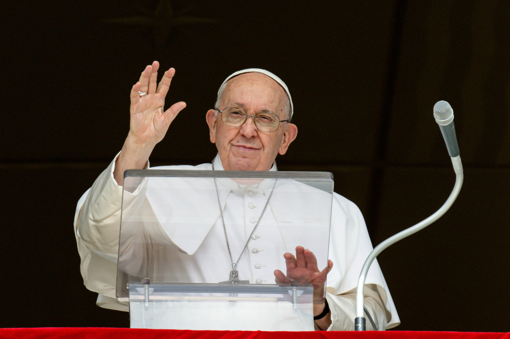 Pope Francis raises his hand to wave with the other hand on his clear lectern. A microphone is next to him.