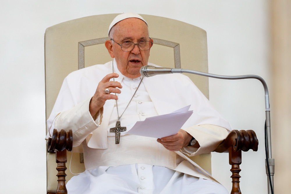 Pope Francis speaks into a microphone while sitting in a large cream chair and holding a piece of paper