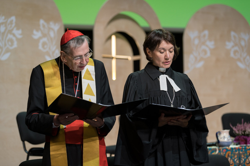 A man wearing a red zucchetto and a yellow stole stands next to a woman wearing a clerical collar and a black robe