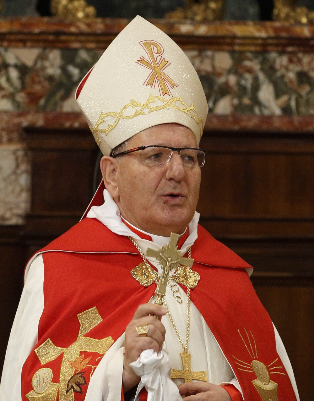 A light-skinned man wears a mitre, glasses, and red and white vestments and carries a gold crucifix