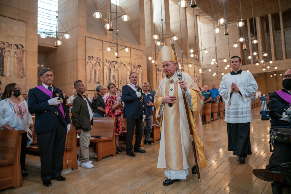 A light-skinned man in golden vestments and a mitre with a crozier walks down a cathedral aisle
