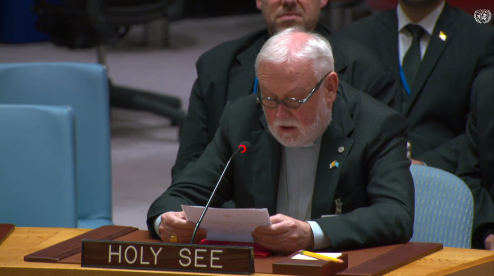 A white-haired white man with a beard and glasses wearing a gray shirt and clerical collar and black suitspeaks into a microphone behind a plaque saying "Holy See"