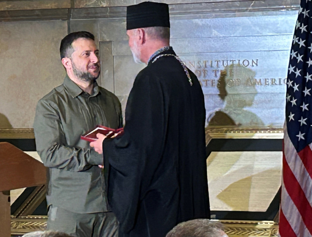 President Volodymyr Zelenskyy, wearing green, hands a red box to a white man wearing a cylindrical black hat and black vestments
