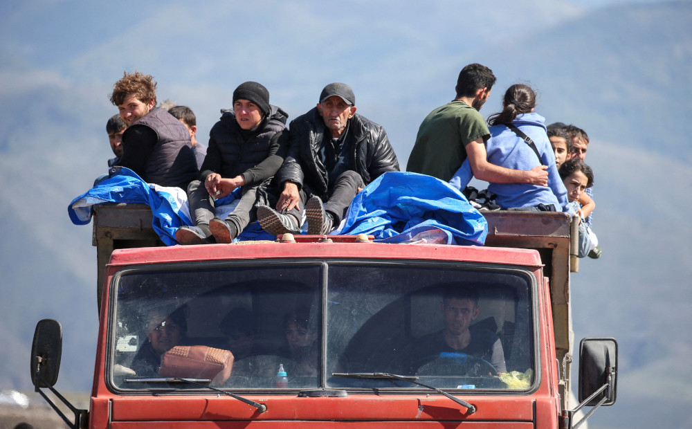 At least ten people sit on the roof of a orangey-red truck. More people are visible in the cab of the truck.