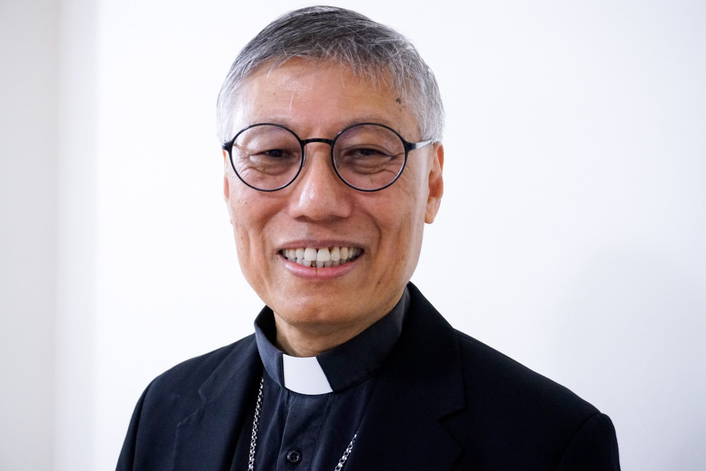 An East Asian man wearing round glasses and a clerical collar smiles at the camera