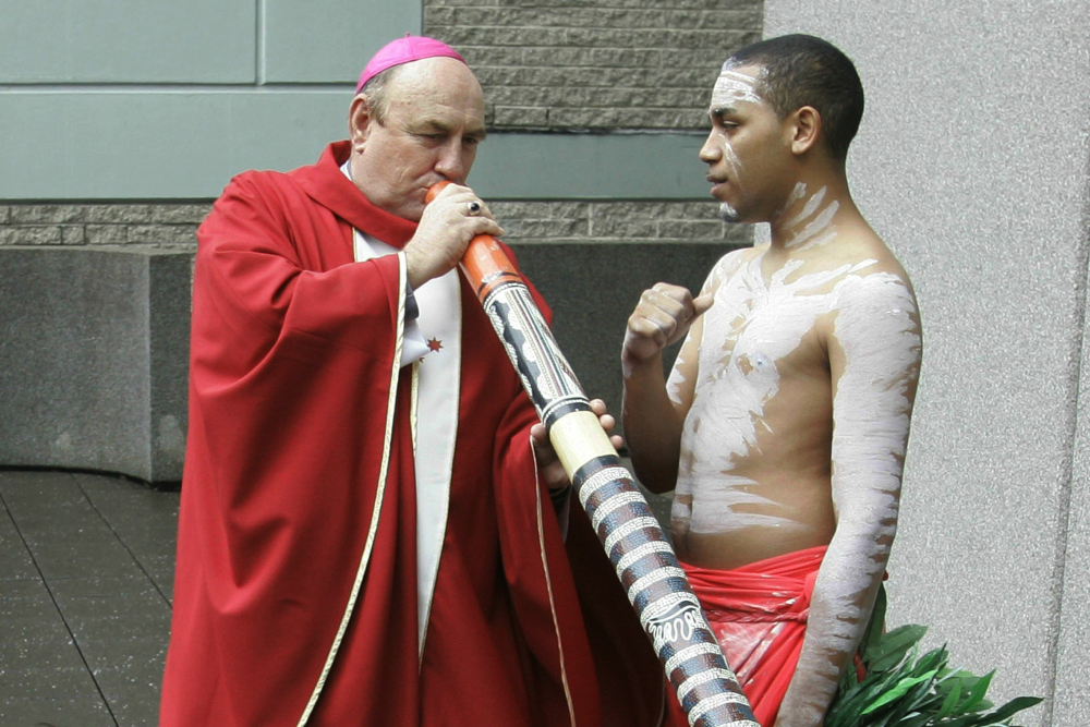 A white man wearing a pink zucchetto and red vestments blows into a digeridoo while standing next to a dark-skinned youth wearing white body paint.