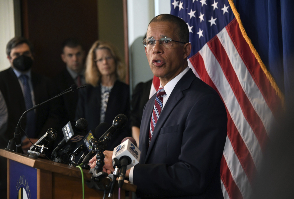 A light-skinned Black man with glasses in a suit speaks from behind a podium with many news microphones