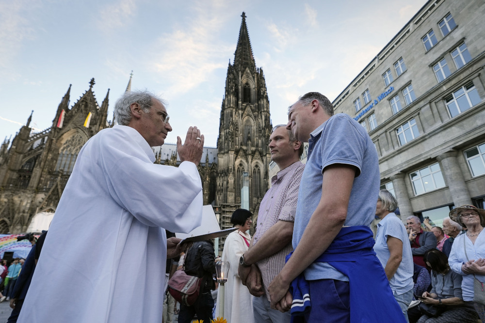 An older white man in white vestments holds his hand as though making the sign of the cross in front of two white men