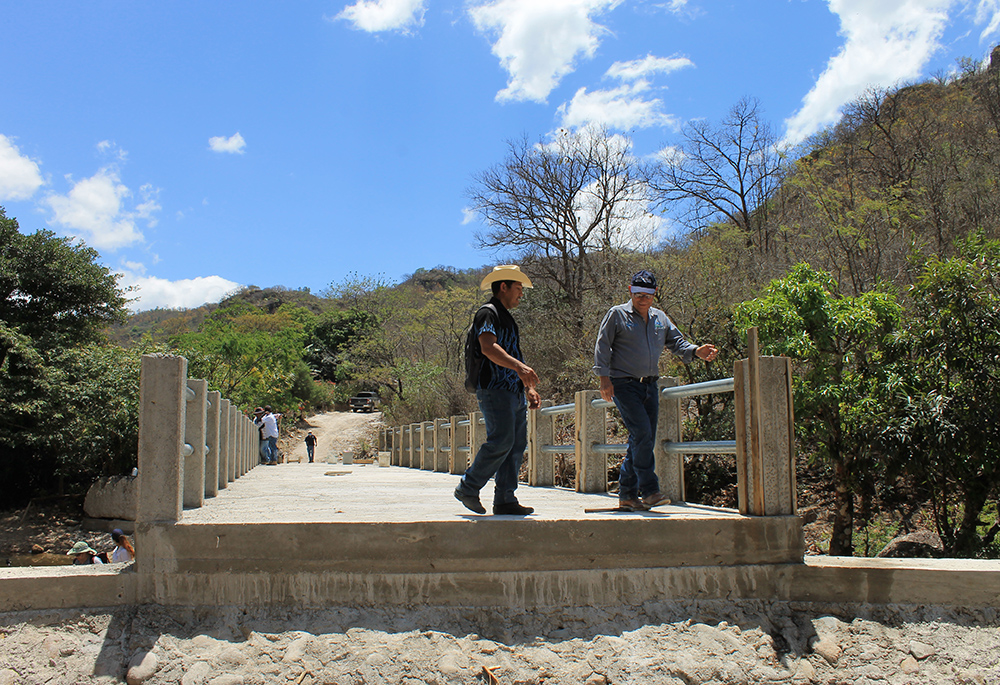 Desiderio Vasquez (cowboy hat), a local farmer, and Miguel Flores (right), a senior technical adviser for Catholic Relief Services Honduras, stand on a bridge being built in the Los Hornos community to connect a long and winding rock-strewn road to a farming community on the other side of a creek. (NCR photo/Brian Roewe)