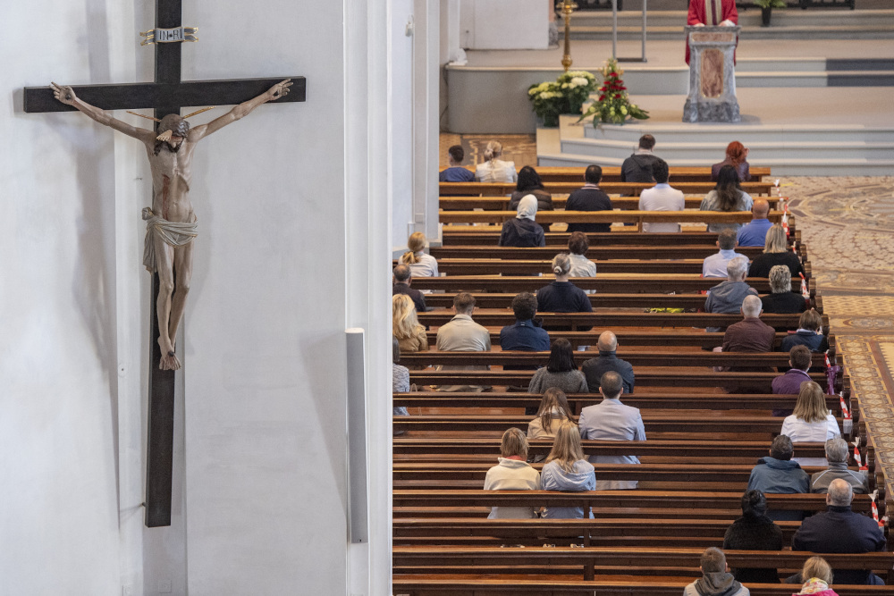 The photo focuses on a crucifix on the wall. People sitting in church pews are also visible