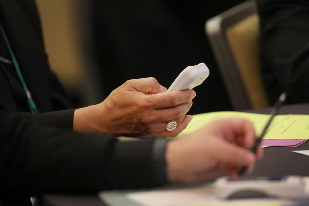 A bishop hands are shown, holding a cellphone size device to cast votes. 