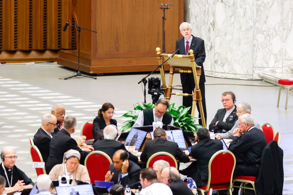 man in a dark suit and red tie stands at lectern and speaks to crowd, A full table of people sits in front of him