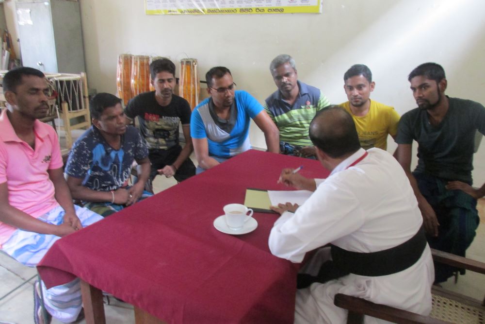 Priest sits behind table as he talks with a group of men.