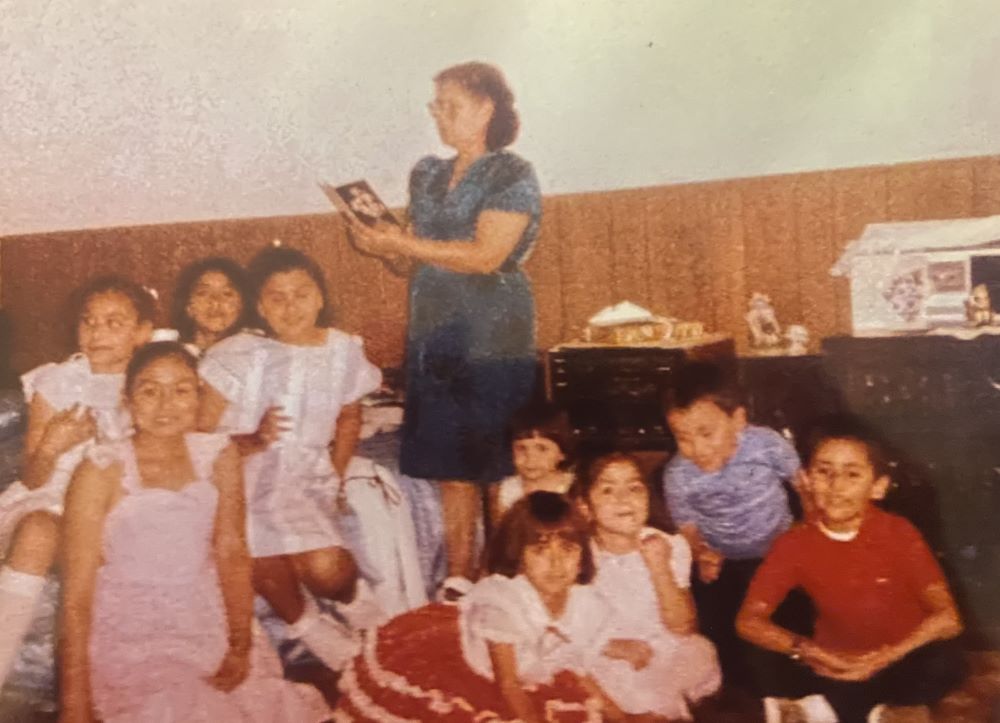 Woman holding book prays with several children seated around her on the floor.