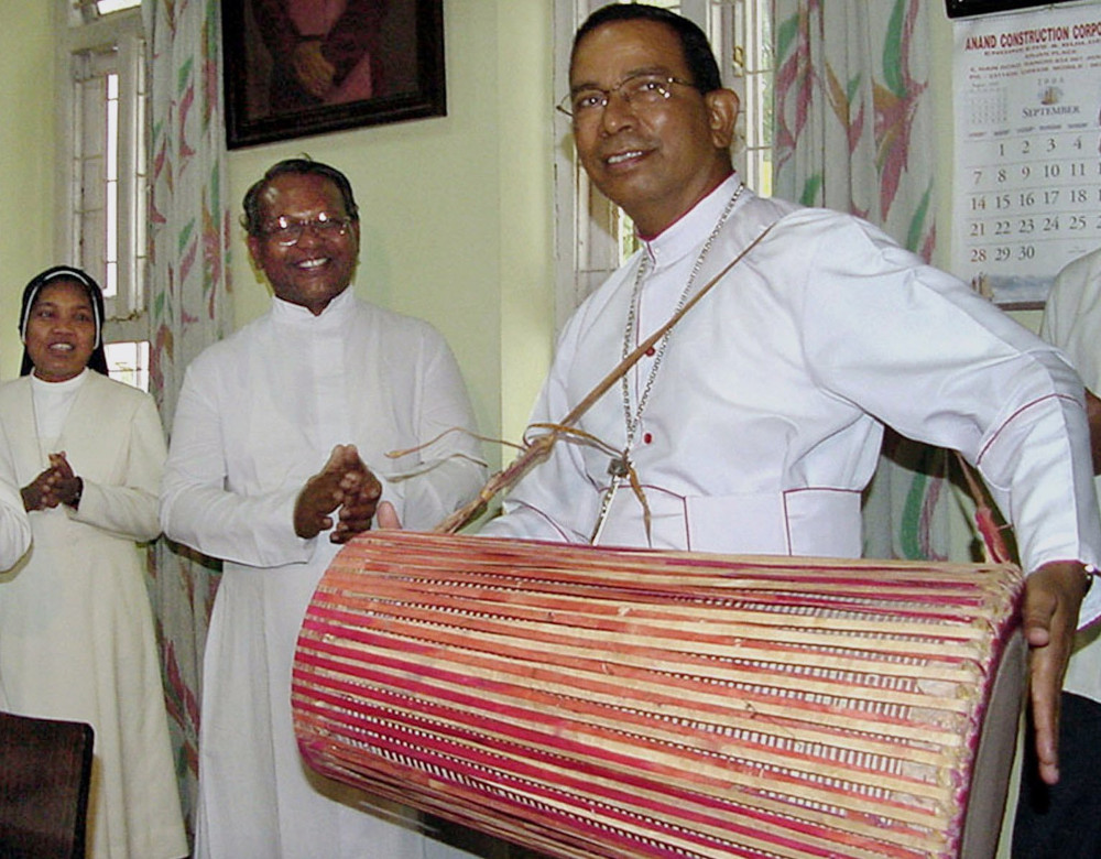 A brown man wearing white has a large drum slung over his shoulder. Another man wearing white and religious sister wearing white stand next to him.