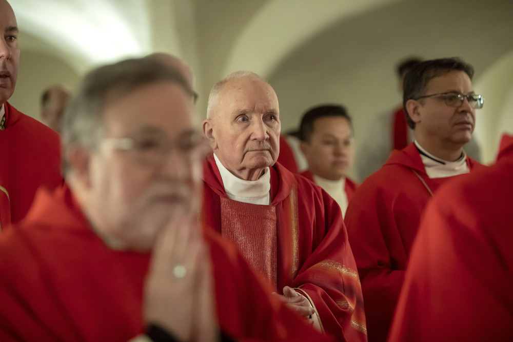 An older white man wearing red vestments stands among other men wearing red vestments