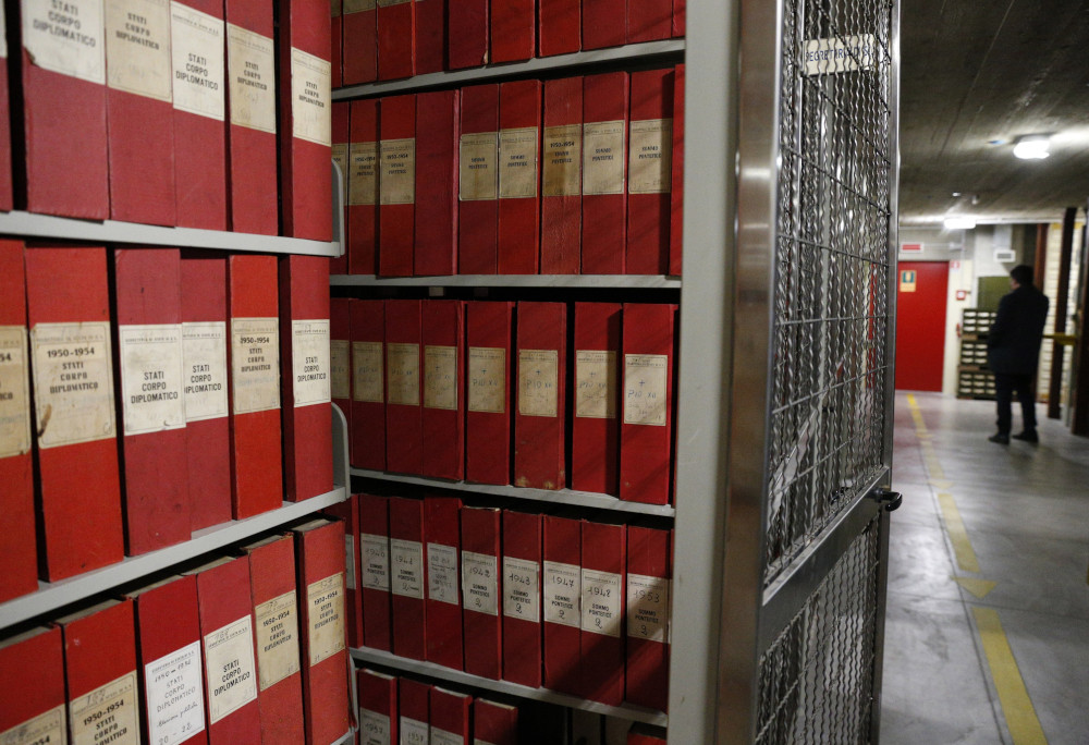 Red binders or upright boxes line metal shelves in a basement. Some have dates on the spines.