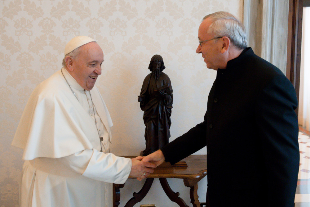 Pope Francis shakes the hand of a white man wearing glasses and a black garment