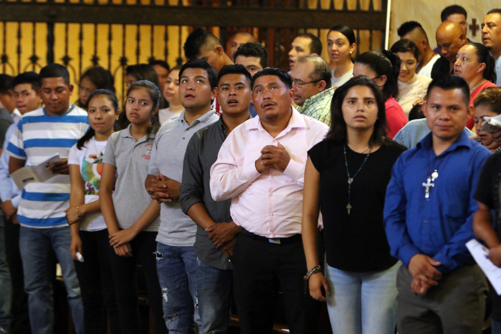 A group of brown people, many wearing crosses, assume prayerful postures