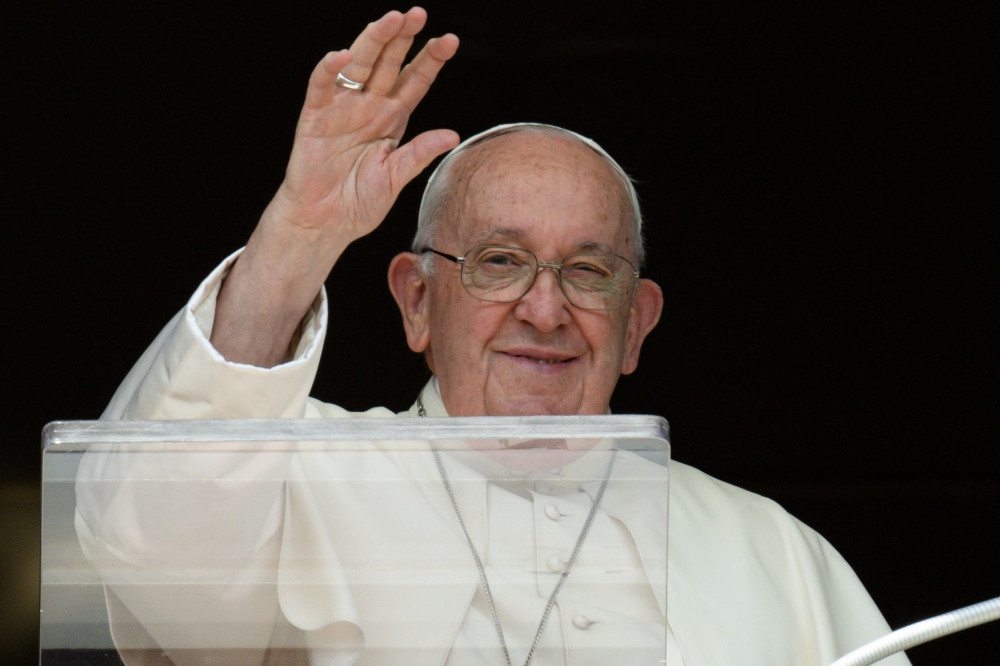 Pope Francis smiles and raises his white hand as he stands behind a clear lectern