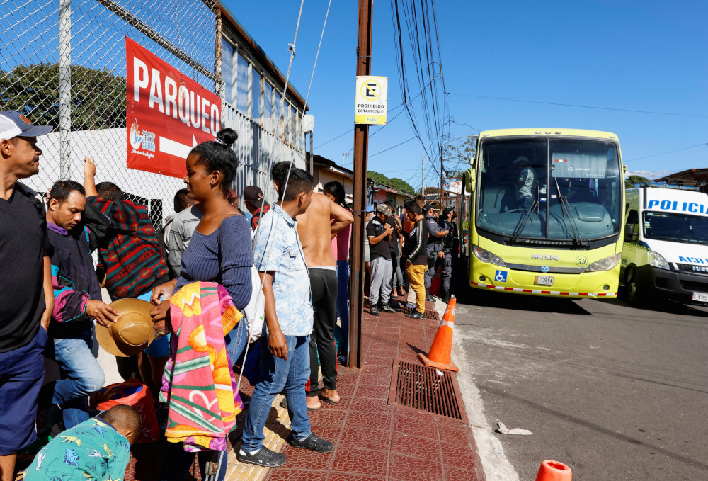 Brown people form a line on a sidewalk in between a chain link fence and a yellow bus