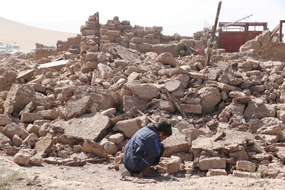 A boy wearing a blue jacket crouches with his face in his hands beside a large pile of rubble