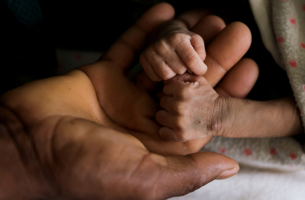 A large brown adult hand holds two incredibly skinny and small brown newborn hands and arms