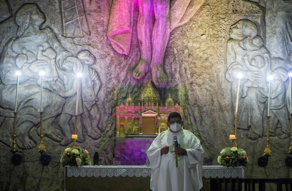 A priest wearing a face mask and white vestments holds a microphone and stands in front of a stone wall with religious carvings and purple light