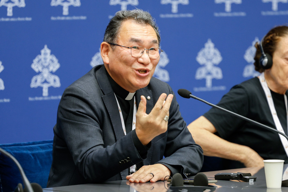A smiling East Asian man wearing glasses and a clerical shirt and jacket speaks into a microphone