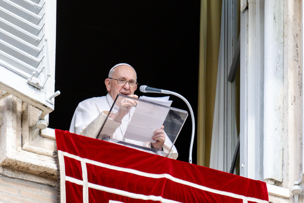 Pope Francis stands in his apartment window and holds a piece of paper above his clear lectern as he speaks into a microphone